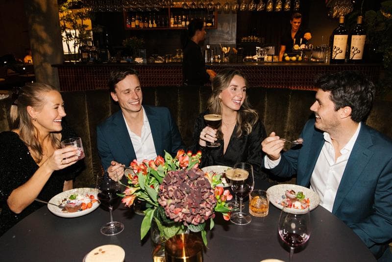 A group of four people sit around a restaurant table, smiling and chatting as they enjoy food and drinks in a warm, atmospheric setting.